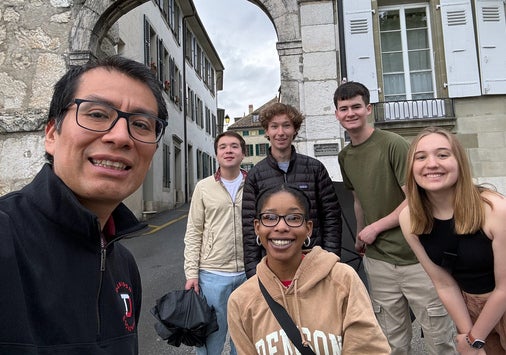 Six people pose and smile for a group photo on a street under a stone archway, with historic buildings in the background.