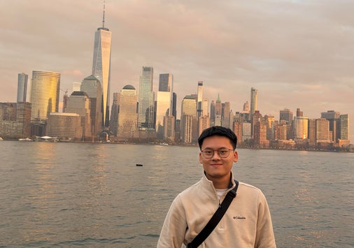 A man wearing glasses and a light jacket stands in front of the New York City skyline by the water at sunset.