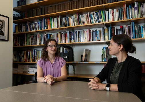 Two women having a conversation at a table in front of a wall of books; a black and white portrait of Denison alum Kirtley Mather hangs on the wall nearby.
