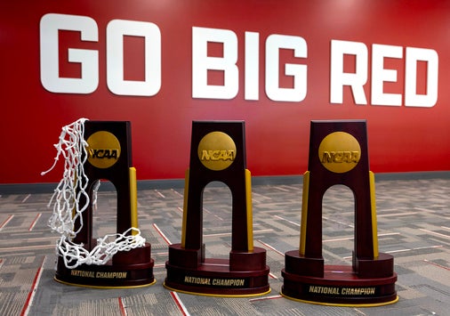 Three NCAA championship trophies lined up in front of a large red well with the white block letters that say "Go Big Red"