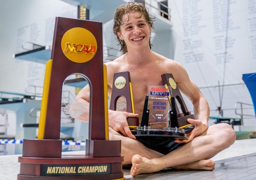 A young man in a swimsuit sits on a pool deck, smiling and holding several NCAA diving trophies, with a large “National Champion” trophy in the foreground and a diving board in the background.