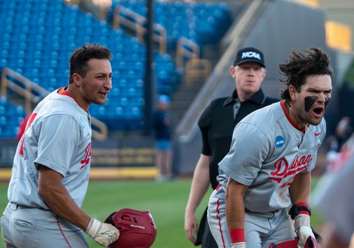 Two baseball players in gray uniforms celebrate on the field; one is holding a helmet and smiling, the other is shouting with black face paint. An NCAA official stands behind them. Blue stadium seats are visible in the background.