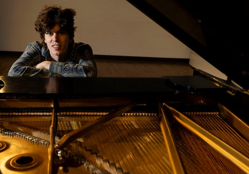 A person with curly hair leans on a grand piano, looking toward the camera. The pianos strings and internal components are visible in the foreground.