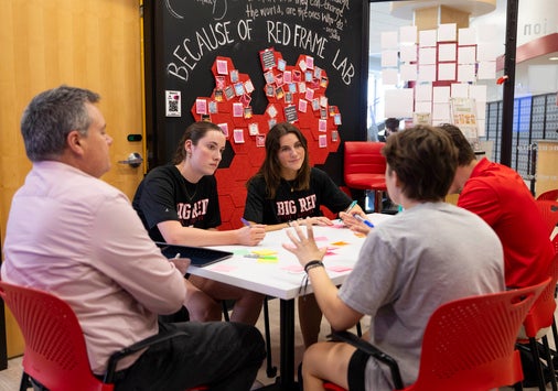 Four people sit around a small white table in discussion, with papers and markers on the table, in a classroom with red chairs and a wall.