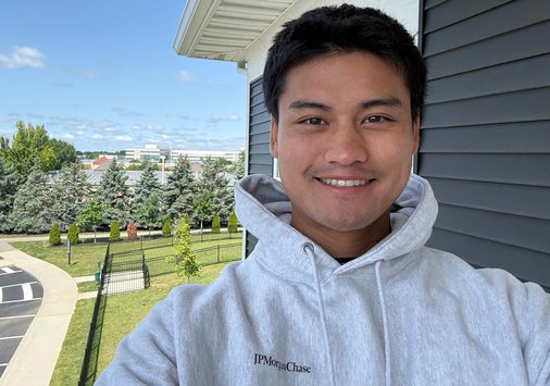 A person wearing a light gray hoodie smiles for a selfie outside near a building, with trees, grass, and a parking lot in the background under a blue sky.