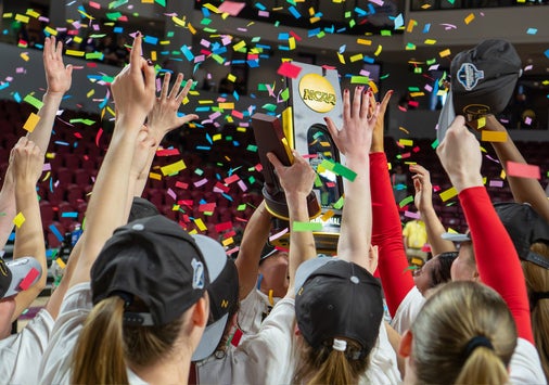 A sports team wearing hats raises a trophy together amid falling confetti in an indoor arena, celebrating a championship victory.