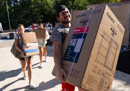 People carry large boxes and belongings outdoors on a sunny day, possibly moving into a dorm or apartment.