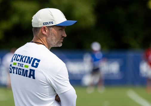 A man in a white Kicking the Stigma shirt and Colts cap stands on a sports field, watching an outdoor practice session.