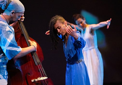 A musician plays an upright bass while two dancers, one in blue and one in white, perform expressive movements on a dimly lit stage.