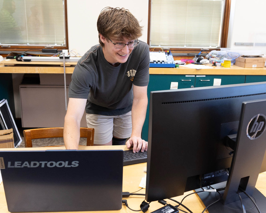 Photo of Denison student Joe Teates ’28 standing at a lab table, looking at computer monitors.