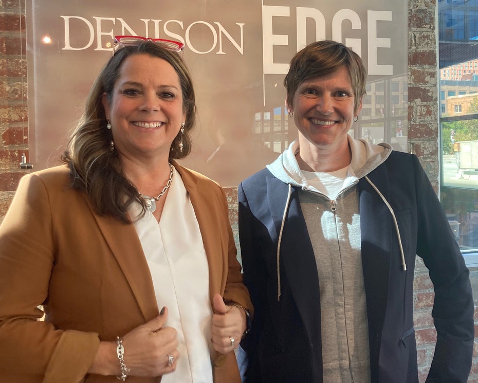 Two people, Kristi Wellington-Baker, Amazon Web Services Higher Education Strategy and Liv Gjestvang, Denison vice president and chief information officer, smiling and posing indoors in front of a sign that reads “DENISON EDGE,” with brick walls and windows in the background.