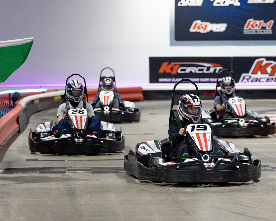 A group of go-karts race on an indoor track as a green flag waves them on.