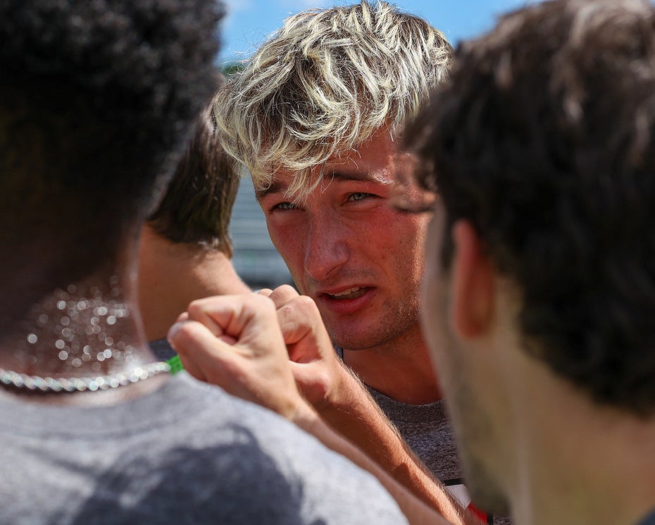 A close up photo of a group of college soccer players on the field; the face of a young man with blond hair is in focus in the center of the photo, surrounded by his teammates giving a celebratory fist bump.