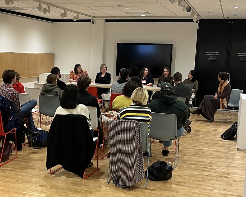 A group of people gathered in a museum gallery listening to a panel discussion. 