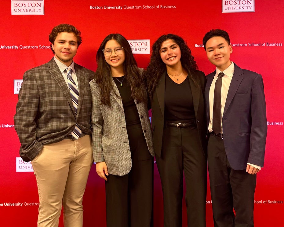 Four Denison students in business attire stand in front of a large red backdrop dotted with Boston University and BU Questrom School of Business logos ahead of their competition.