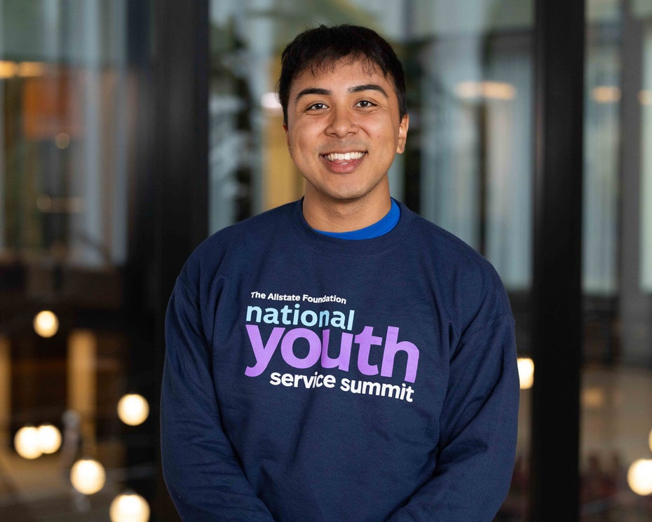 Photo of Yahir Fernandez-Alvarez ’26 wearing a National Youth Service Summit shirt, standing in front of a large glass wall inside a modern building.