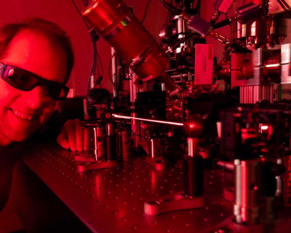 A man in dark protective glasses smiles next to a scientific piece of equipment emitting a laser in a red-hued room.