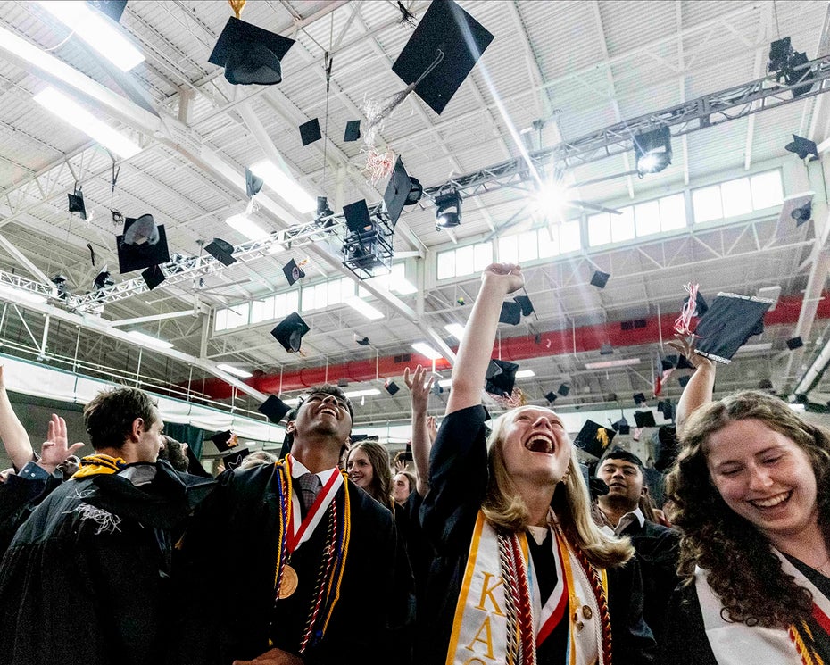 A large group of college graduates in caps and gowns toss their caps in the air in celebration in a large fieldhouse during a graduation ceremony.