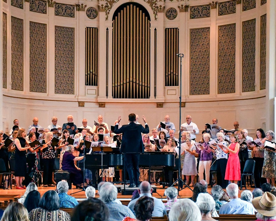 A choir and musicians perform on stage in a church or concert hall, directed by a conductor. The audience is seated in rows facing the performers. The background features a large organ and decorative architectural details.