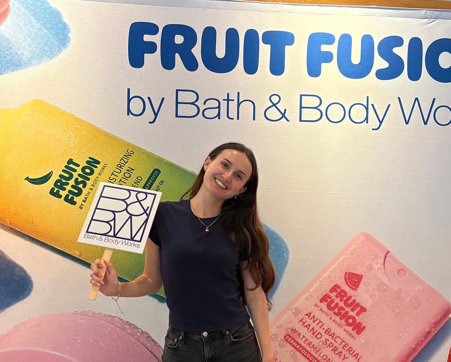 A person stands in front of a Bath & Body Works Fruit Fusion display, holding a sign and smiling at the camera.
