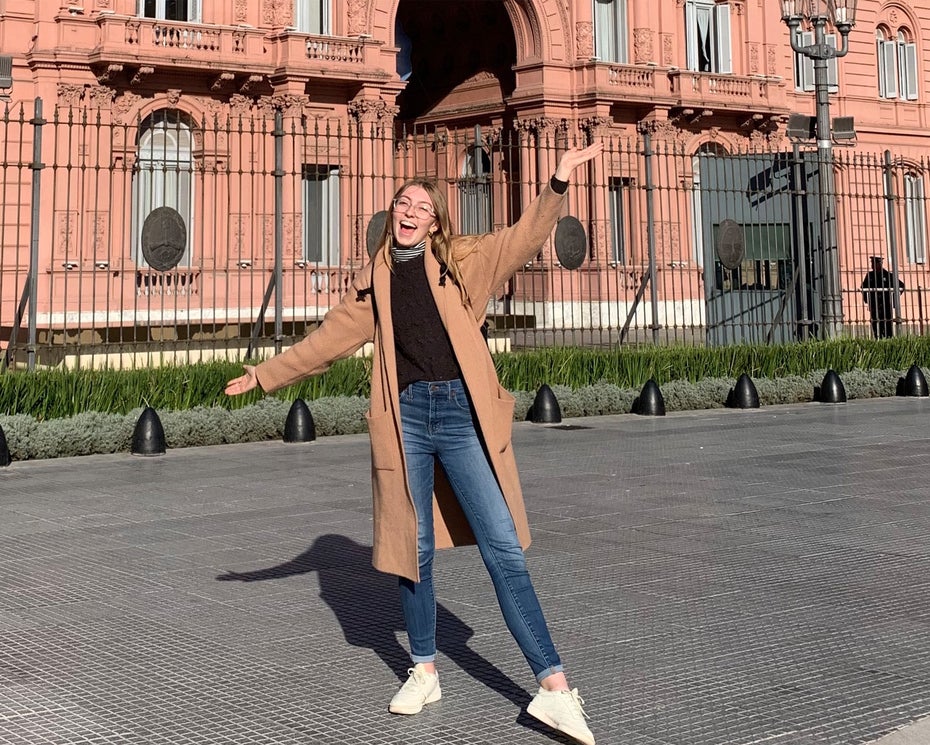 A study abroad student in a tan coat and jeans stands smiling with arms outstretched in front of a large pink historic building with black iron fencing.