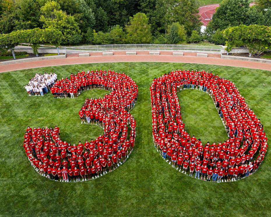 A large group of people wearing red shirts stand on grass arranged to form the number 30, with a few people in white shirts forming a dot before the 3. Trees and a path are in the background.