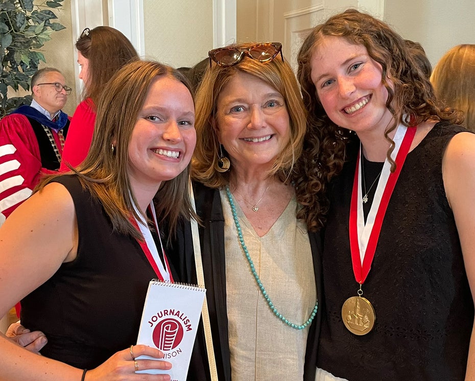 Three women stand indoors, smiling at the camera. Two younger women wear medals on red and white ribbons around their necks, and the other woman stands between them. Other people are visible in the background.