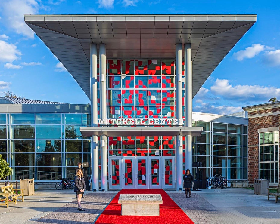 Front view of the Mitchell Recreation & Athletic Center main entrance with large glass windows, red accents, and a red carpet leading to the entrance under a modern overhang.
