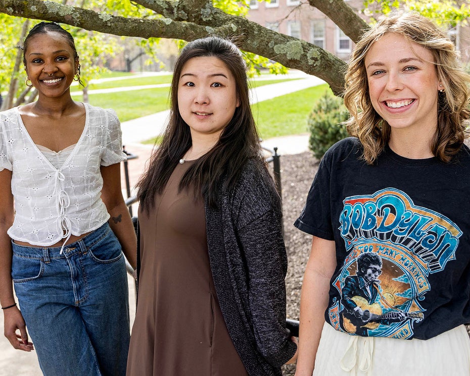 Three young women stand outdoors on a sunny day, posing and smiling in front of a leafy tree and campus buildings.