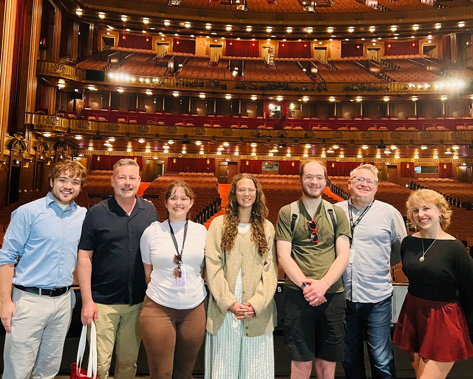 A group of seven people stand on a theater stage with rows of empty red seats and ornate balconies visible behind them.
