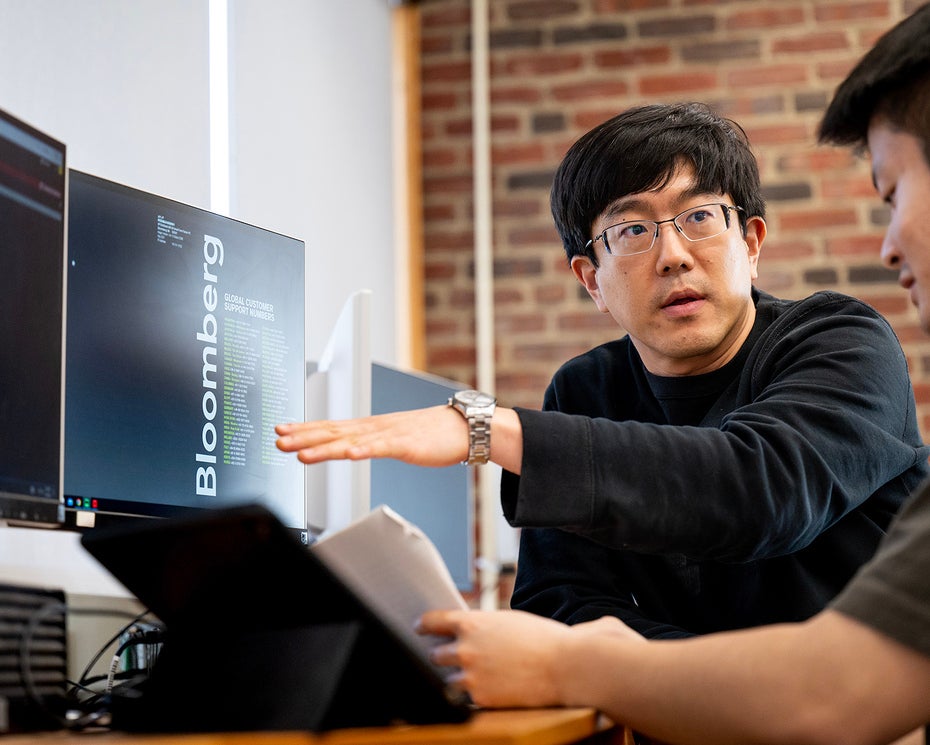 Two people work at a desk with computer monitors and a tablet, discussing data displayed on the screens in a modern office with exposed brick walls.
