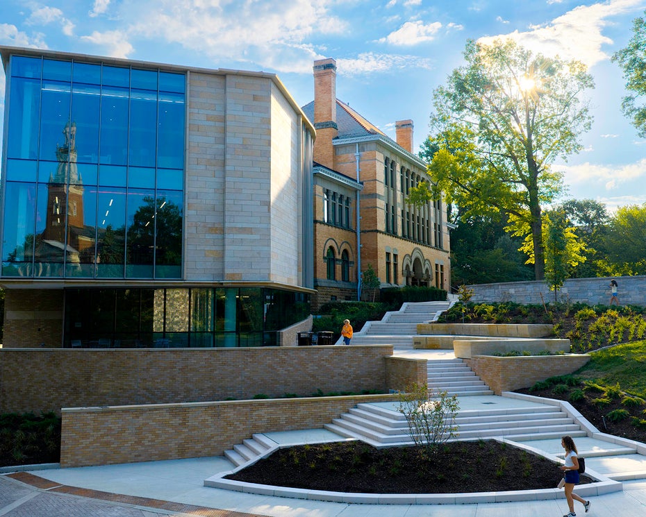Modern glass and brick building with large windows, outdoor steps, and landscaped greenery under a partly cloudy sky.