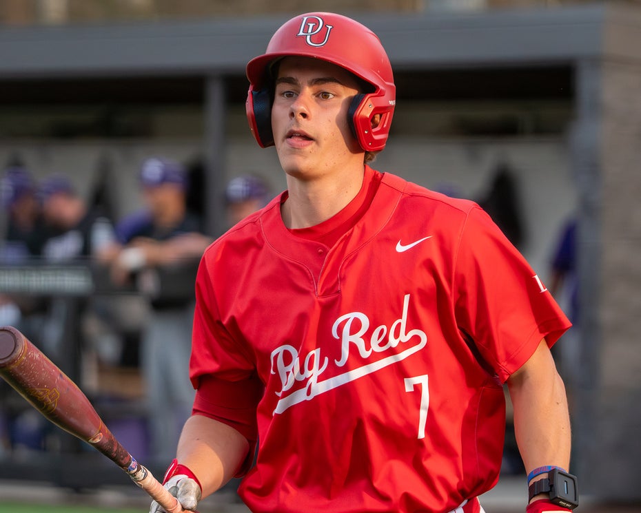 Baseball player in a red uniform and helmet holding a bat stands near the field, with a dugout and other players visible in the background.