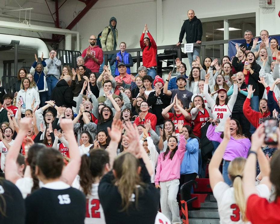 A crowd of fans fill the bleachers as women's basketball players salute them from the court.