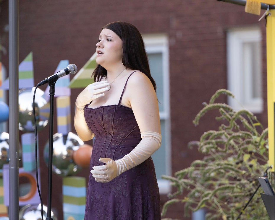 A woman with dark hair, wearing a purple sleeveless dress with straps, sings on an outdoor stage.