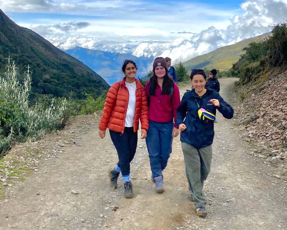 Three people on a hiking on a high-altitude trail with snow-capped mountains and clouds in the distance behind them.