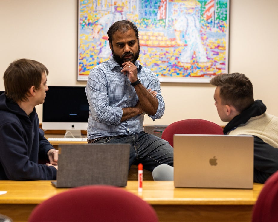 A man sits on a desk speaking with two students in a classroom with computers and colorful artwork on the wall in the background.