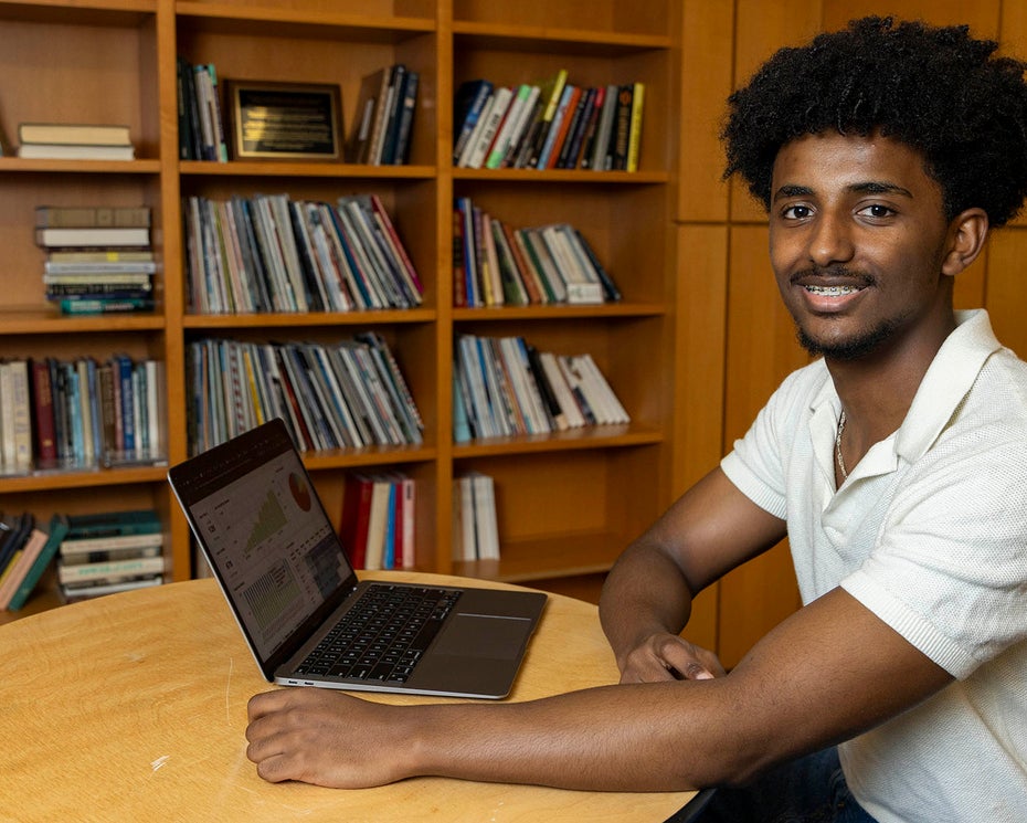 A person sits at a round table in front of a laptop, smiling, with bookshelves filled with books in the background.