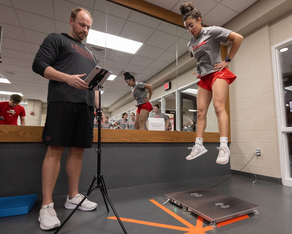 A woman in athletic gear performs a vertical jump test while a man in sportswear records results on a tablet. The scene takes place in a gym with mirrors and exercise equipment visible in the background.