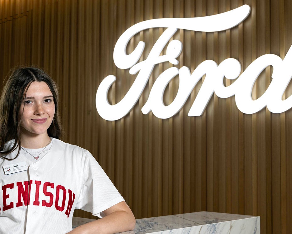 A person in a Denison t-shirt stands in front of a large illuminated Ford logo on a wood-paneled wall.