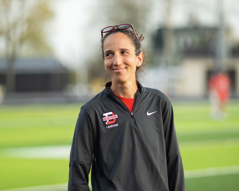 A woman with dark hair in athletic gear stands outside on a sports field.