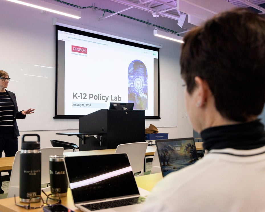 A person stands and presents in front of a projected slide titled "K-12 Policy Lab" to an audience seated with laptops in a classroom.