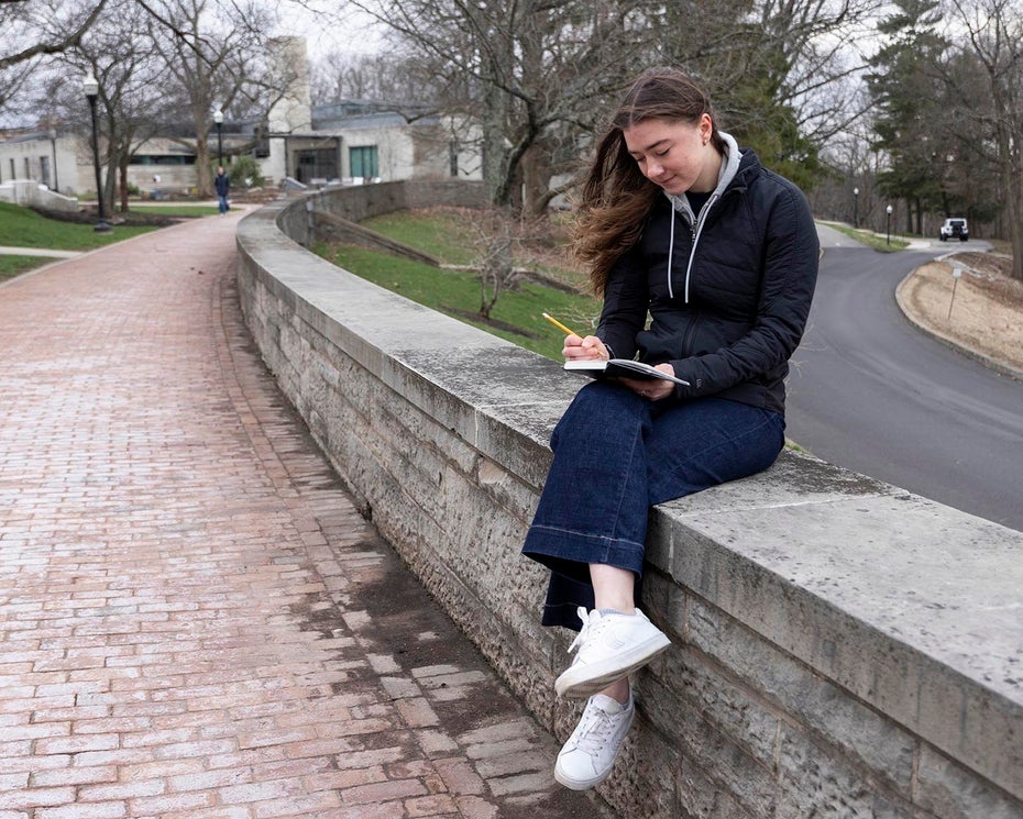 A young woman sits on a curved stone wall along a brick path writing in her notebook.