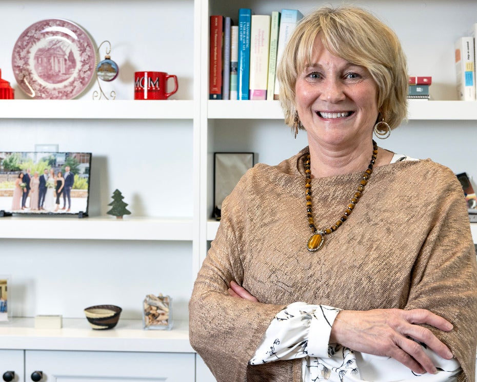A smiling woman with short blonde hair stands in front of white bookshelves filled with books and decorative items, wearing a textured brown top, a necklace, and earrings, with her arms crossed.