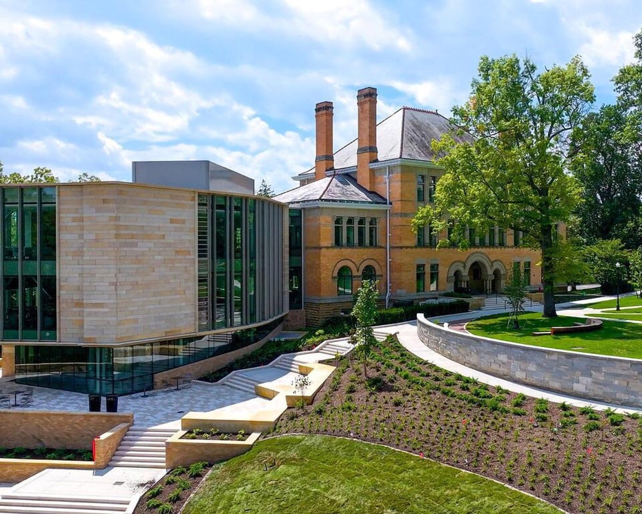 A modern building extension with glass and stone sits beside a large historic brick house, surrounded by green lawns, trees, and landscaped pathways under a partly cloudy sky.