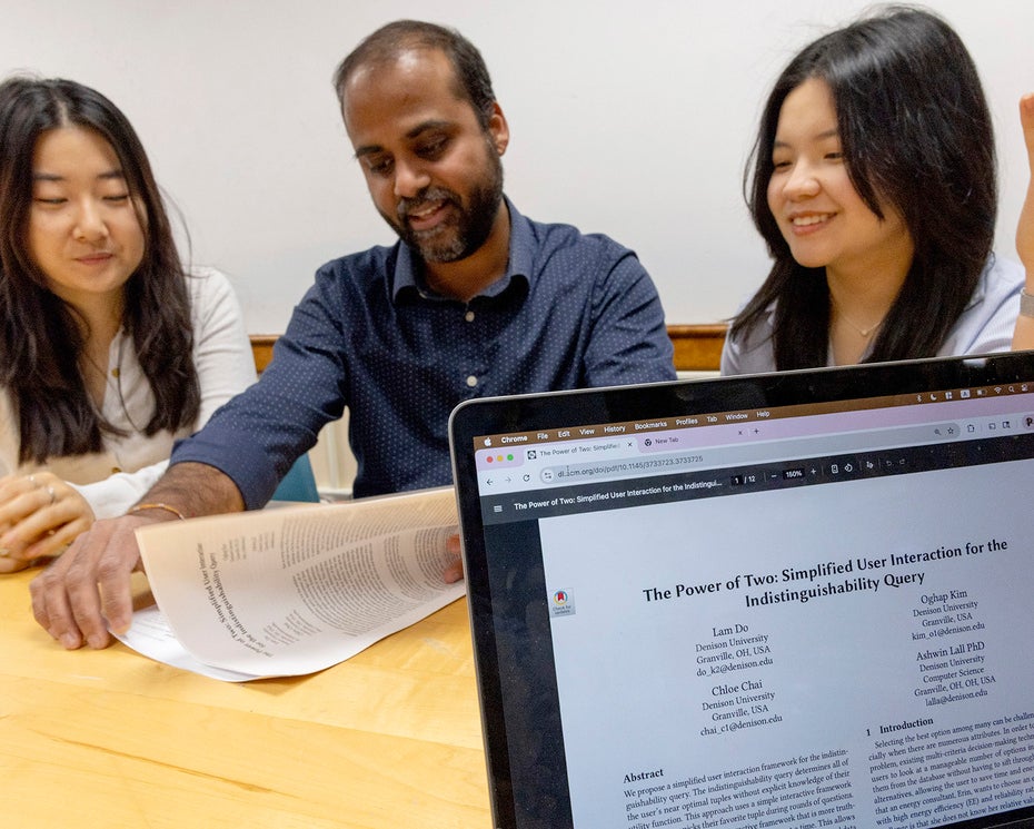 Two college students and a faculty member sit at a table discussing a printed document, which is digitally presented on a laptop screen in the foreground.