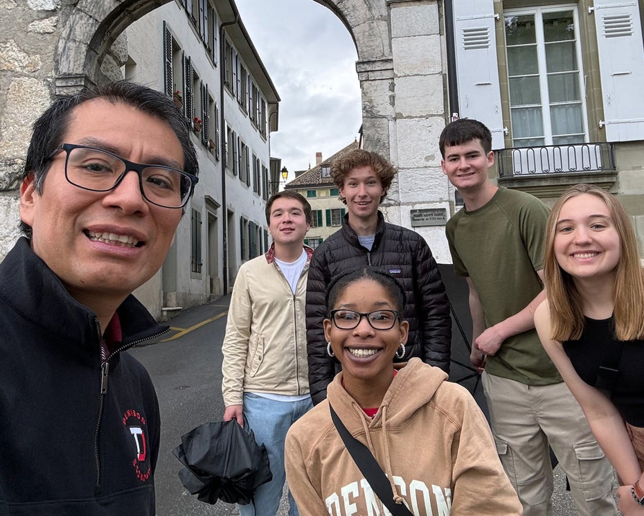 Six people pose and smile for a group photo on a street under a stone archway, with historic buildings in the background.