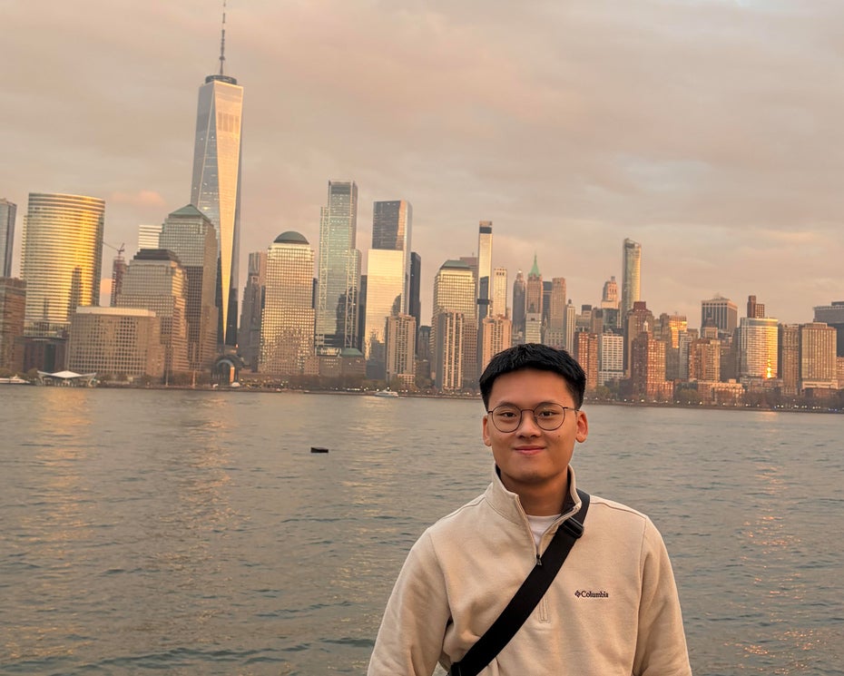 A man wearing glasses and a light jacket stands in front of the New York City skyline by the water at sunset.