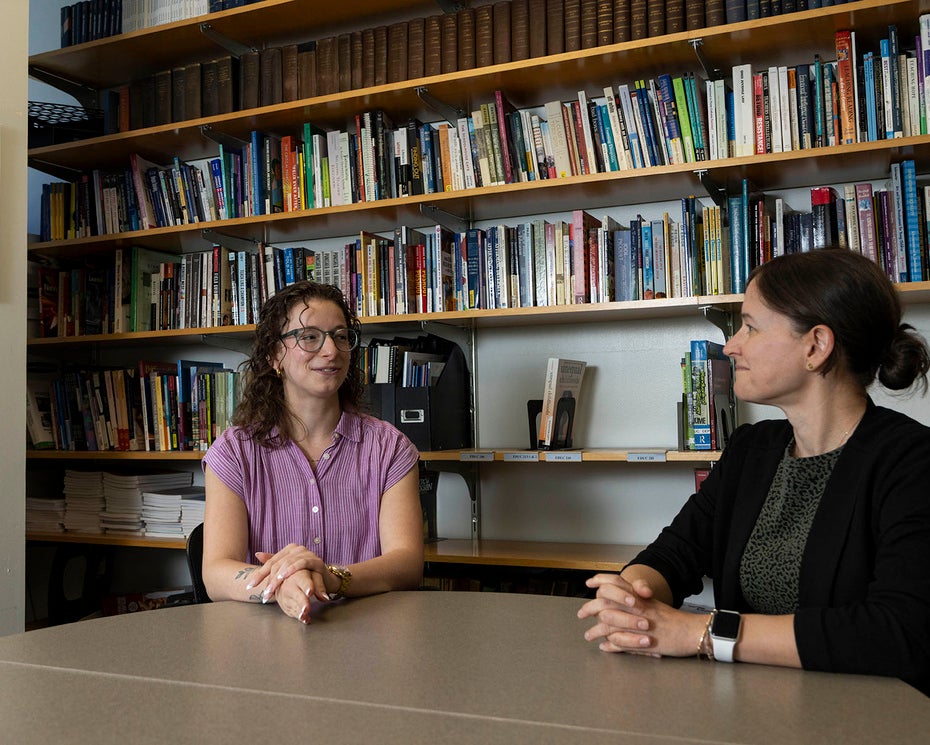 Two women having a conversation at a table in front of a wall of books; a black and white portrait of Denison alum Kirtley Mather hangs on the wall nearby.