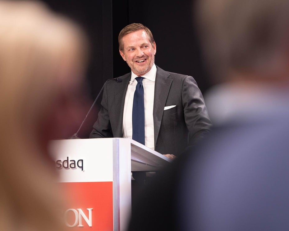 A man in a suit and tie stands at a podium, speaking into a microphone, with a smile on his face. The foreground is blurred, showing parts of two people facing him.